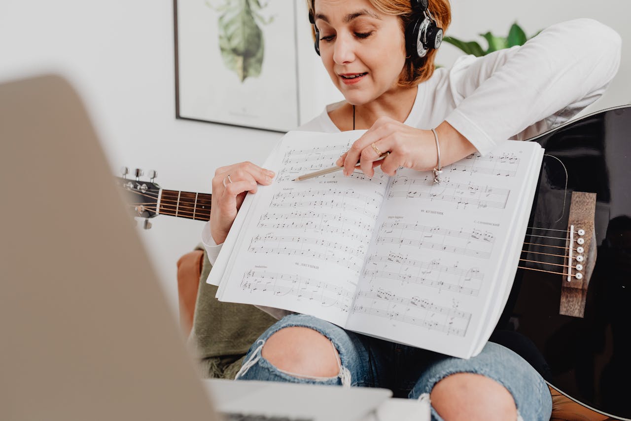A woman teaching a music lesson online, holding a guitar and music sheet, wearing headphones.